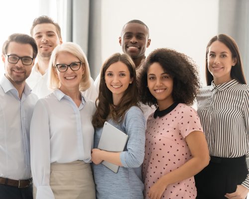 Multiethnic smiling businesspeople standing looking at camera making group photo in office together, happy diverse employees posing for picture with boss or team leader, showing unity and support